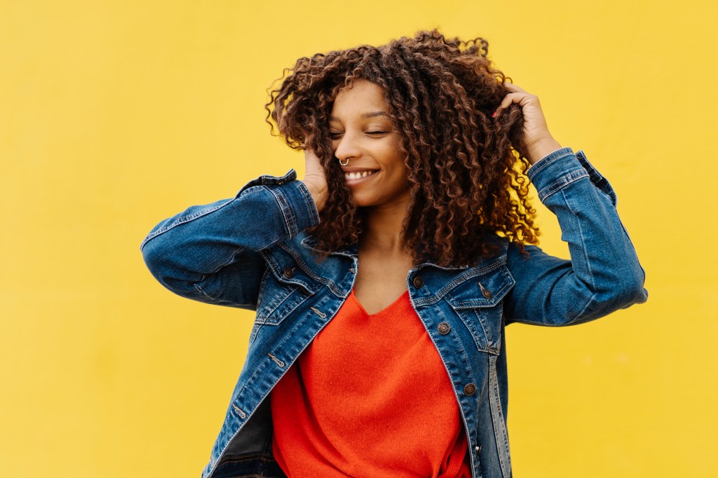 Young woman with African roots and curly hair posing and smiling – Stock photo Mulher com cabelo cacheado mexendo no mesmo enquanto sorri