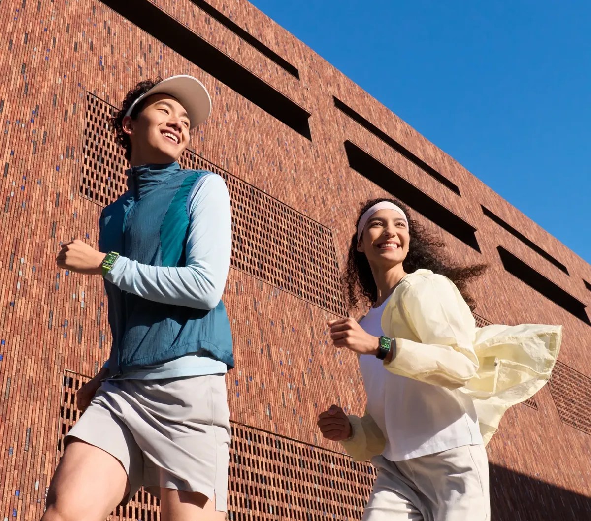 Um homem e uma mulher sorrindo, correndo ao ar livre sob um céu azul, com um prédio de tijolos ao fundo. Ambos usam relógios inteligentes e roupas esportivas leves, o vento movendo a jaqueta da mulher