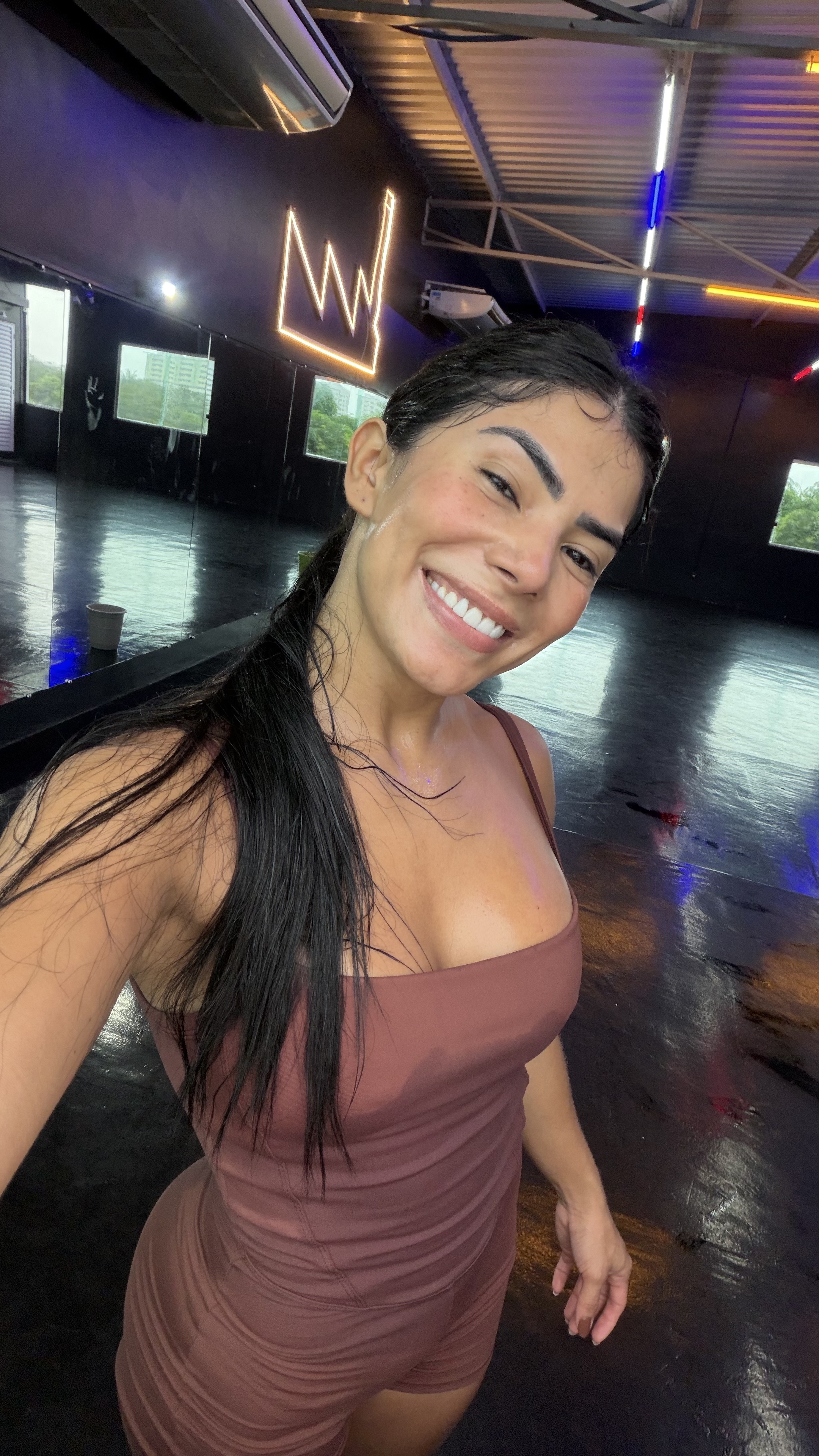 Mulher sorridente, cabelo escuro preso, vestindo um macac&atilde;o marrom, suada ap&oacute;s exerc&iacute;cio. Ela est&aacute; em um est&uacute;dio com piso escuro e reflexivo, paredes pretas, janelas e um letreiro de neon em forma de coroa na parede.