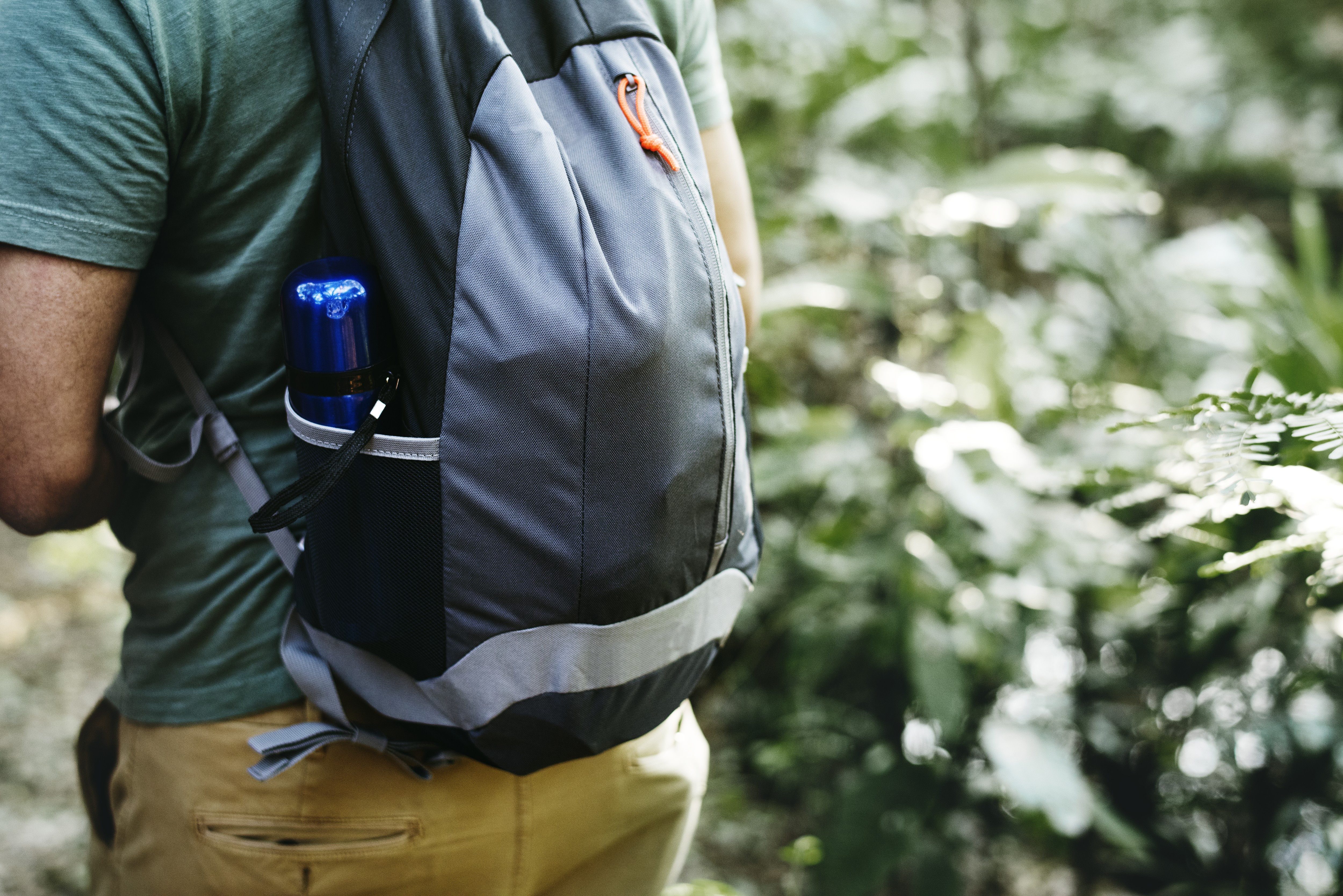 Pessoa de costas, usando camiseta verde-oliva e calça cáqui, com uma mochila cinza e preta. Uma garrafa d'água azul está no bolso lateral da mochila, em meio a folhagens verdes e luz solar