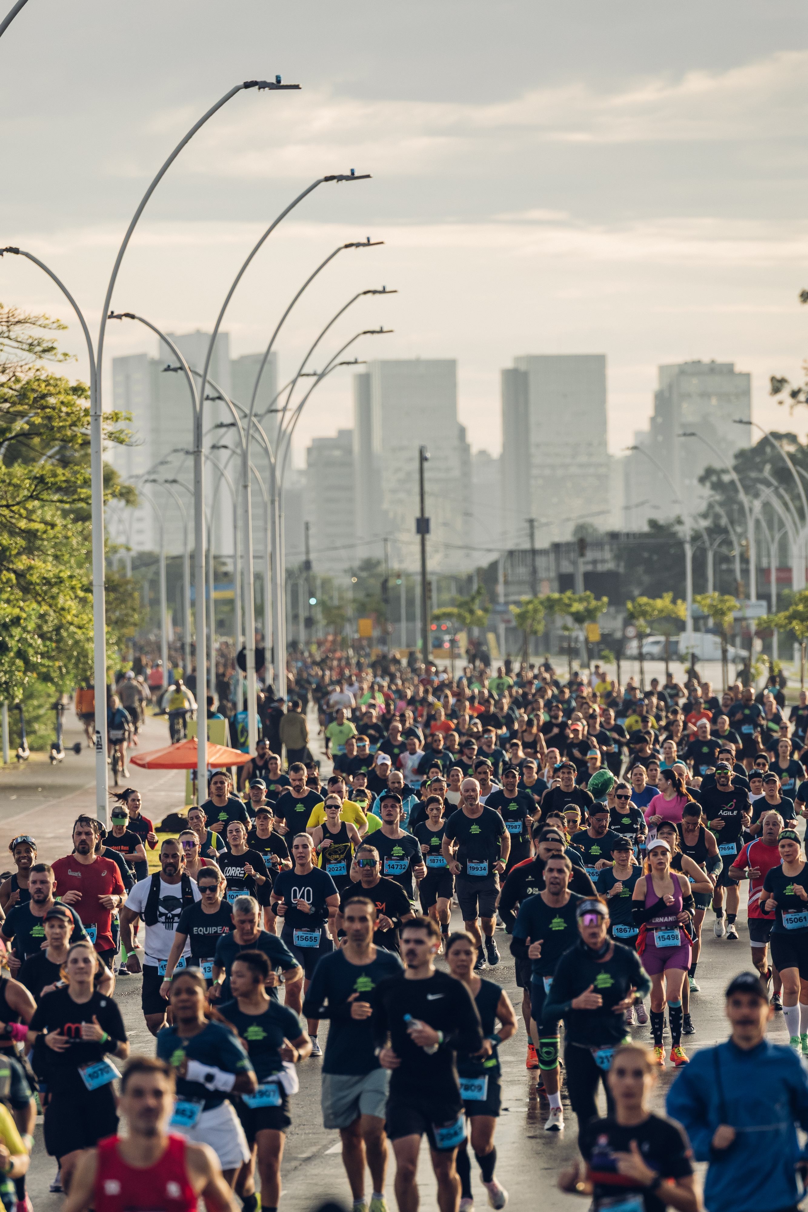 Multid&atilde;o de corredores em uma maratona urbana, vestindo camisetas escuras e n&uacute;meros de peito, em uma rua larga com postes de luz curvos e pr&eacute;dios altos ao fundo, sob c&eacute;u nublado