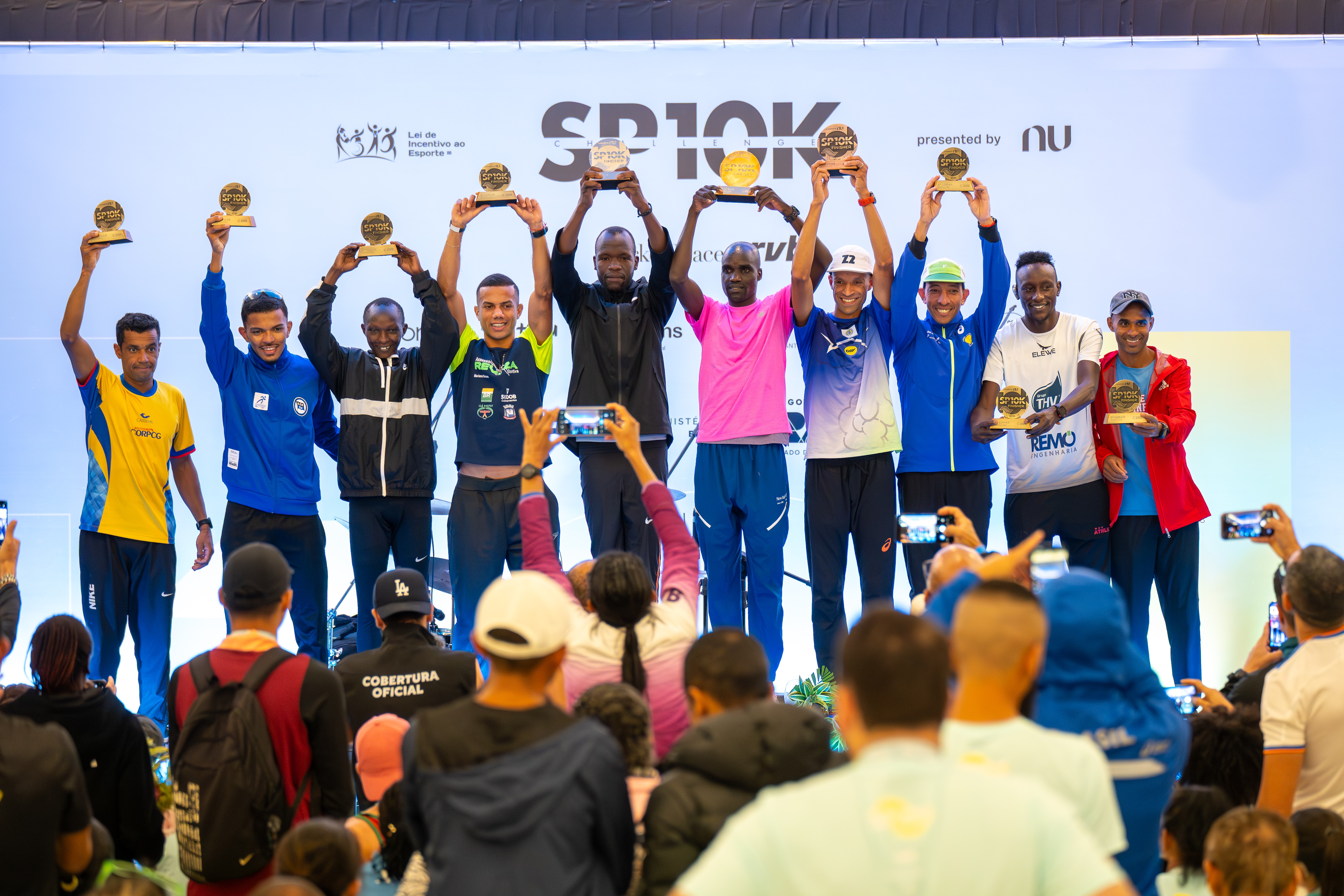 Dez corredores masculinos sorridentes, de diversas etnias, erguem trof&eacute;us dourados no p&oacute;dio da corrida SP10K, com um p&uacute;blico os fotografando