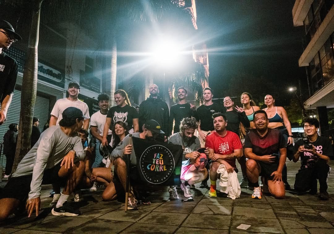 Grupo diverso de cerca de vinte pessoas, algumas agachadas e outras em p&eacute;, posando para foto noturna em rua iluminada por forte luz de poste. Um homem agachado segura uma bandeira preta com a frase FAZ TEU CORRE em letras coloridas. Todos sorriem, vestindo roupas esportivas, indicando um coletivo de corrida.