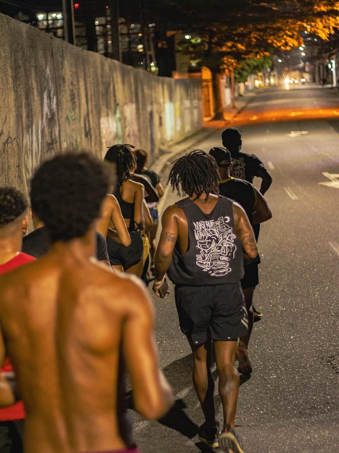 Grupo de pessoas correndo &agrave; noite em uma rua escura, com um muro de concreto &agrave; esquerda e luzes de postes e carros ao fundo. O corredor em primeiro plano est&aacute; sem camisa, e outro usa regata preta com estampa branca nas costas