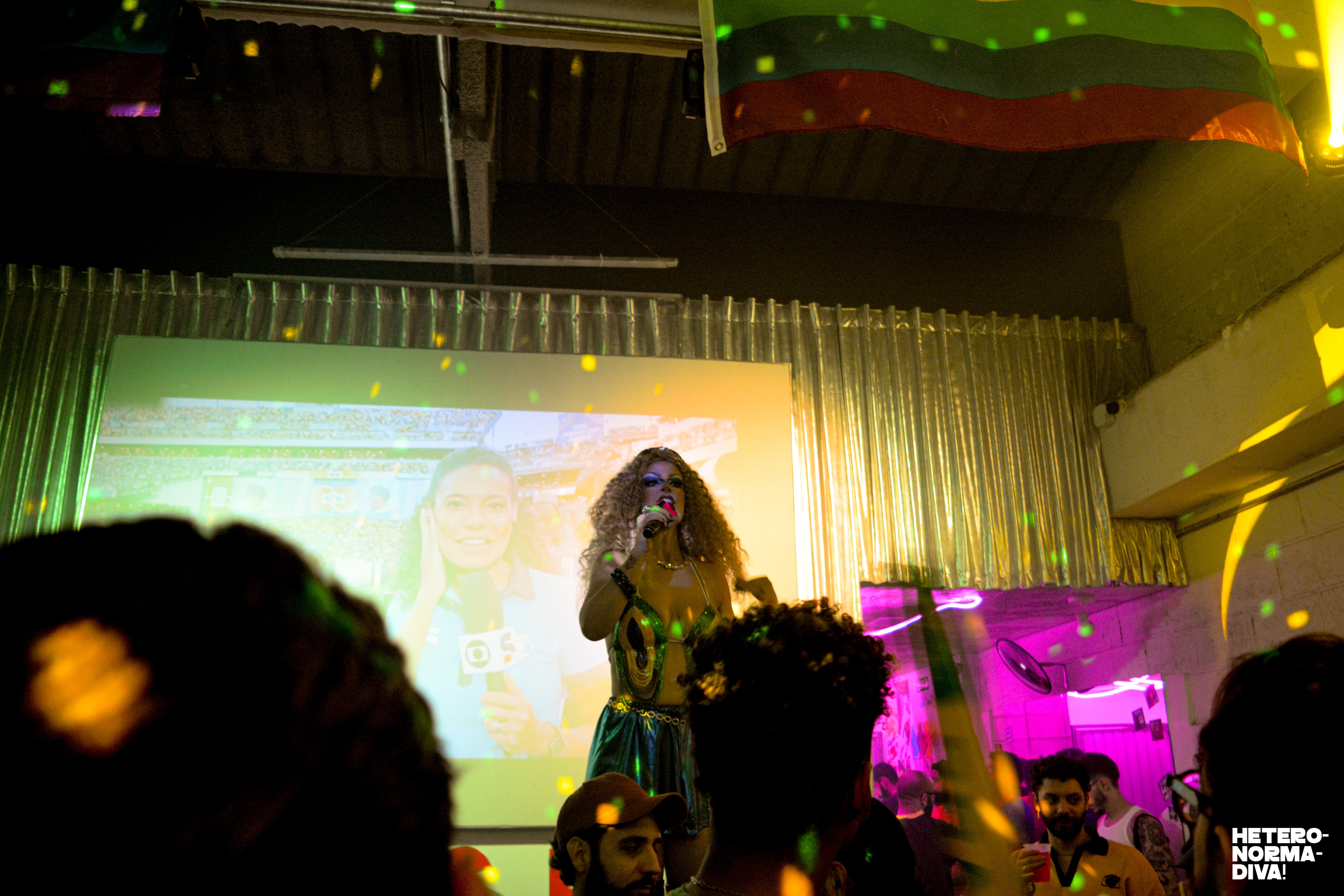 Uma drag queen com cabelo cacheado e maquiagem forte canta no palco, vestindo top dourado e saia verde, com um microfone na m&atilde;o. Atr&aacute;s dela, um tel&atilde;o exibe uma rep&oacute;rter da TV Globo. Pontos de luz coloridos iluminam o ambiente, onde pessoas assistem ao show. Uma bandeira verde, amarela e vermelha est&aacute; pendurada no teto