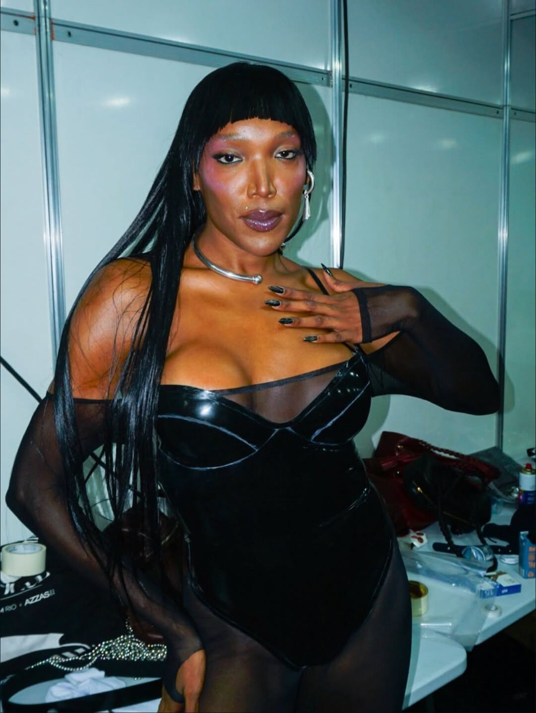 Pessoa de pele escura, cabelo liso preto com franja, usando body de l&aacute;tex preto e mangas transparentes, colar prateado e brincos longos. Ela tem maquiagem roxa nos olhos e l&aacute;bios, unhas pretas e a m&atilde;o esquerda no peito. O fundo &eacute; um camarim com uma mesa bagun&ccedil;ada