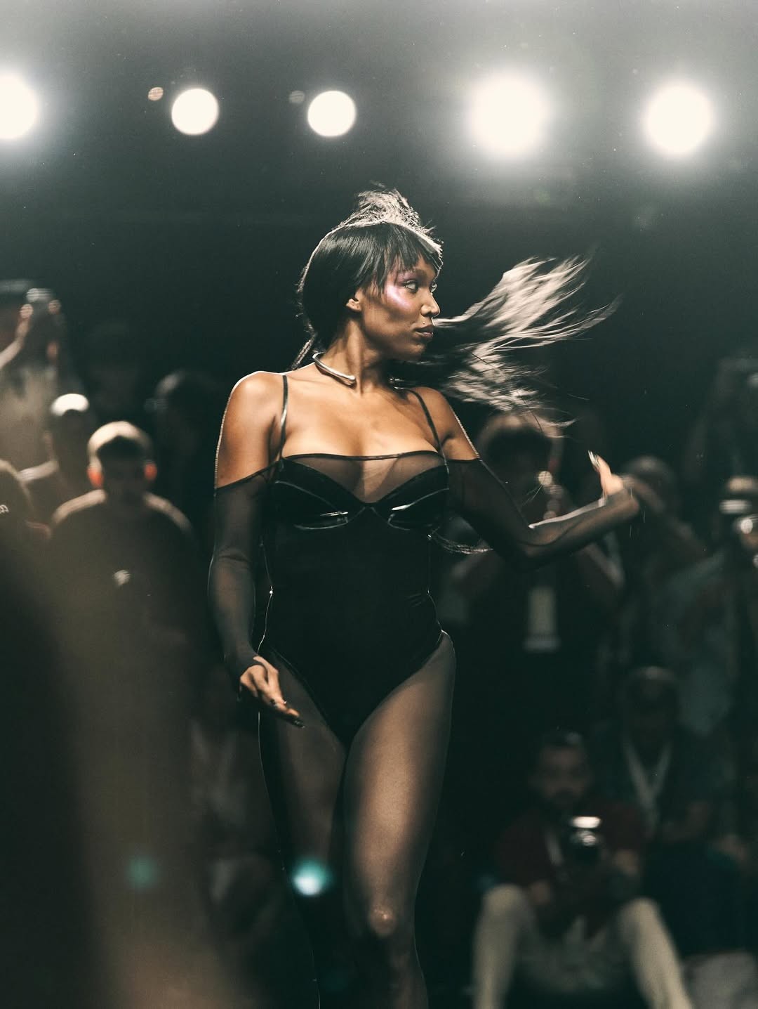 Modelo de pele escura com cabelo esvoa&ccedil;ante, usando body preto transparente e gargantilha prateada, desfilando em passarela escura com luzes fortes e p&uacute;blico desfocado ao fundo