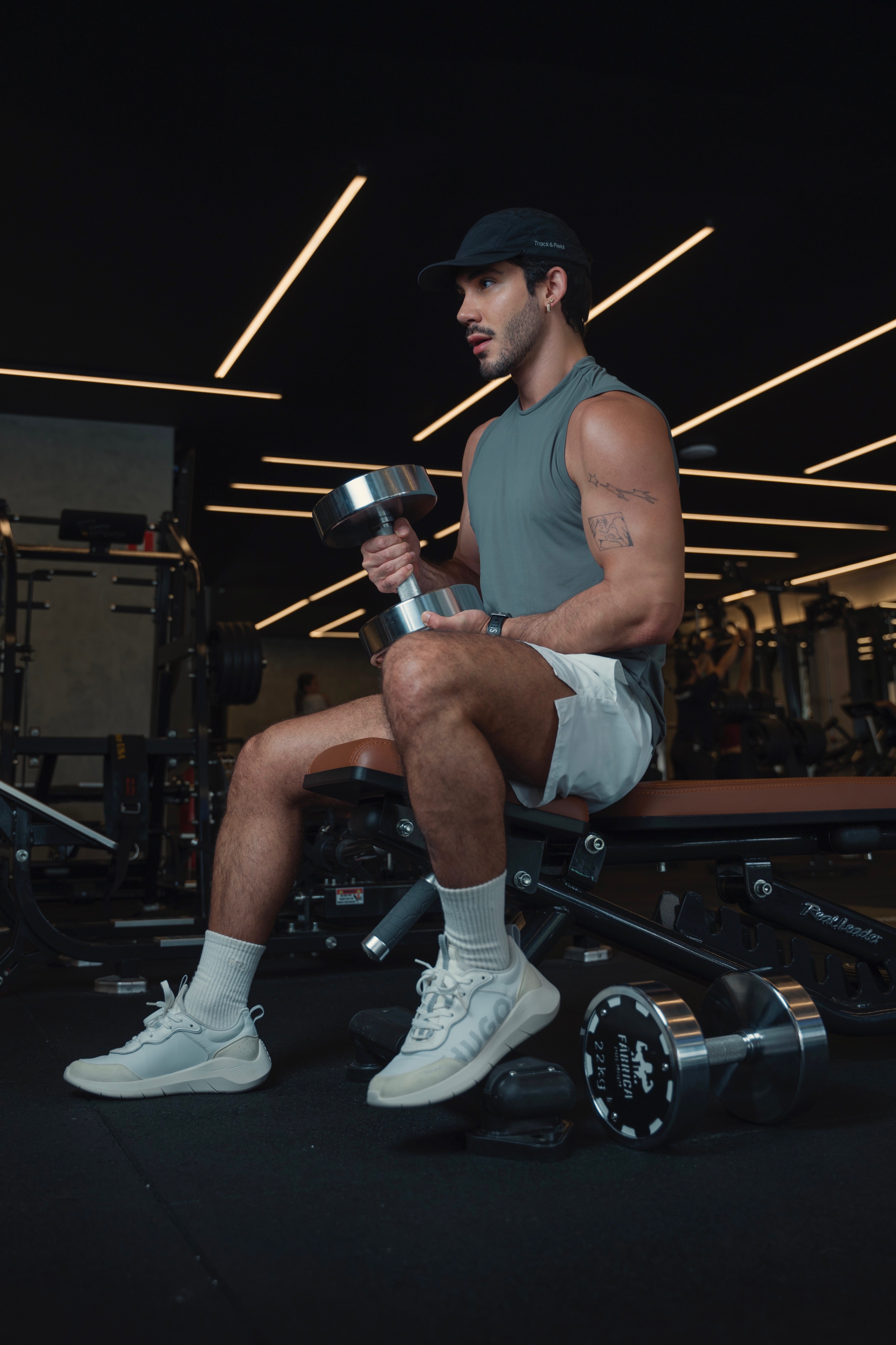 Homem jovem, de bon&eacute; preto e regata cinza, sentado em um banco de academia, segurando um halter prateado com a m&atilde;o direita, olhando para a direita. Ele usa shorts brancos, meias e t&ecirc;nis claros, e tem uma tatuagem no bra&ccedil;o esquerdo. O ambiente &eacute; uma academia com ilumina&ccedil;&atilde;o de neon no teto e equipamentos de muscula&ccedil;&atilde;o ao fundo