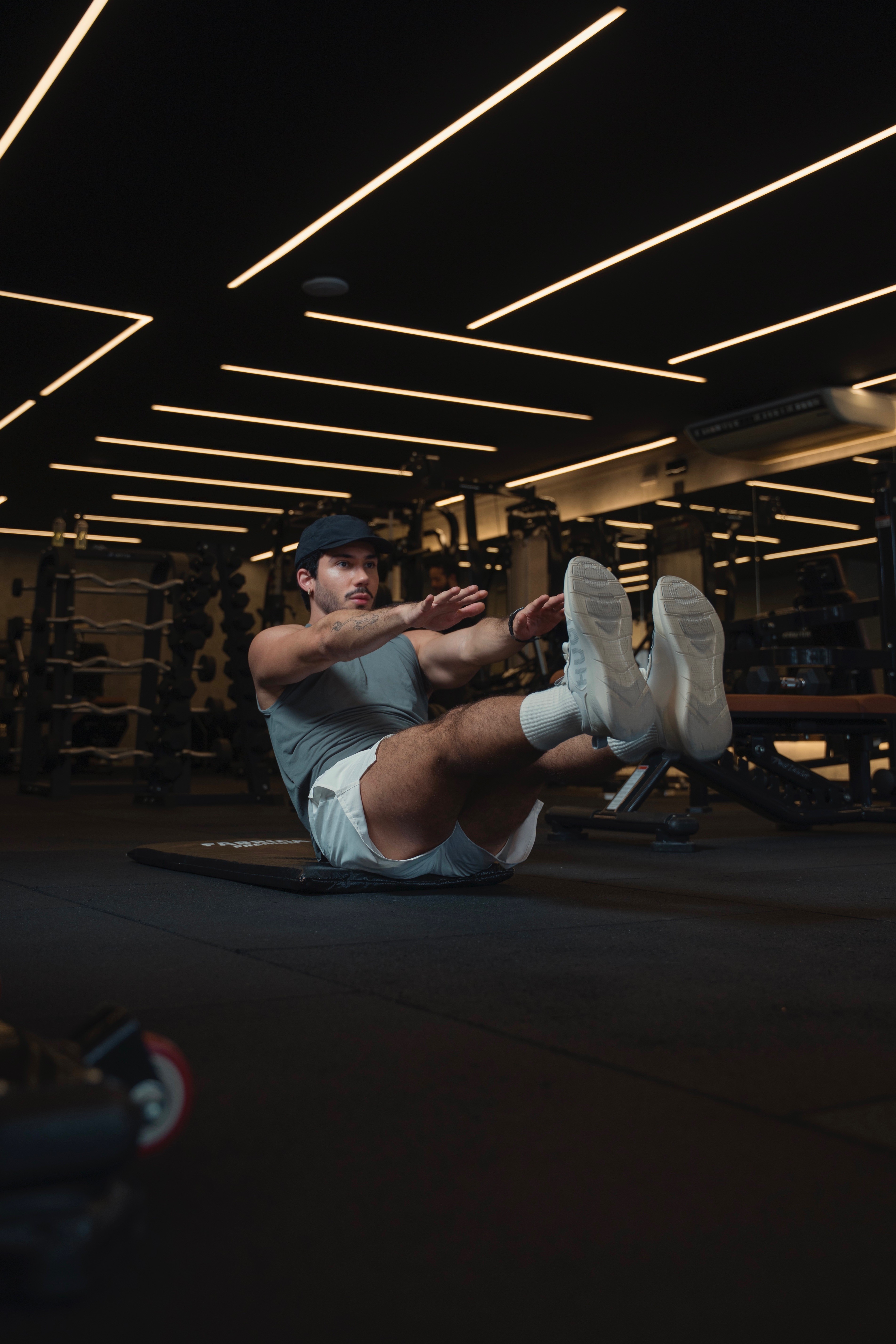 Homem jovem com bermuda e sapatos brancos, camiseta cinza e usando bon&eacute; em uma academia, fazendo abdominal com os bra&ccedil;os esticados.