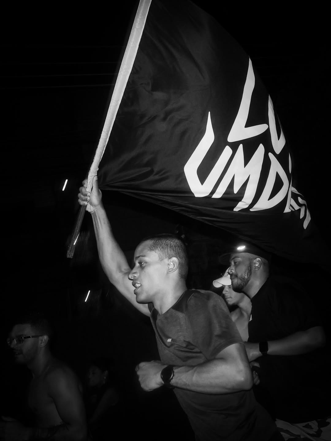 Homem jovem com cabelo curto e rabo de cavalo, vestindo camiseta escura, ergue uma bandeira preta com letras brancas LUMD em um protesto noturno. Ele tem express&atilde;o s&eacute;ria e est&aacute; cercado por outras pessoas, uma delas com bon&eacute; e barba, em segundo plano. A imagem em preto e branco tem ilumina&ccedil;&atilde;o pontual, criando um clima de mobiliza&ccedil;&atilde;o.