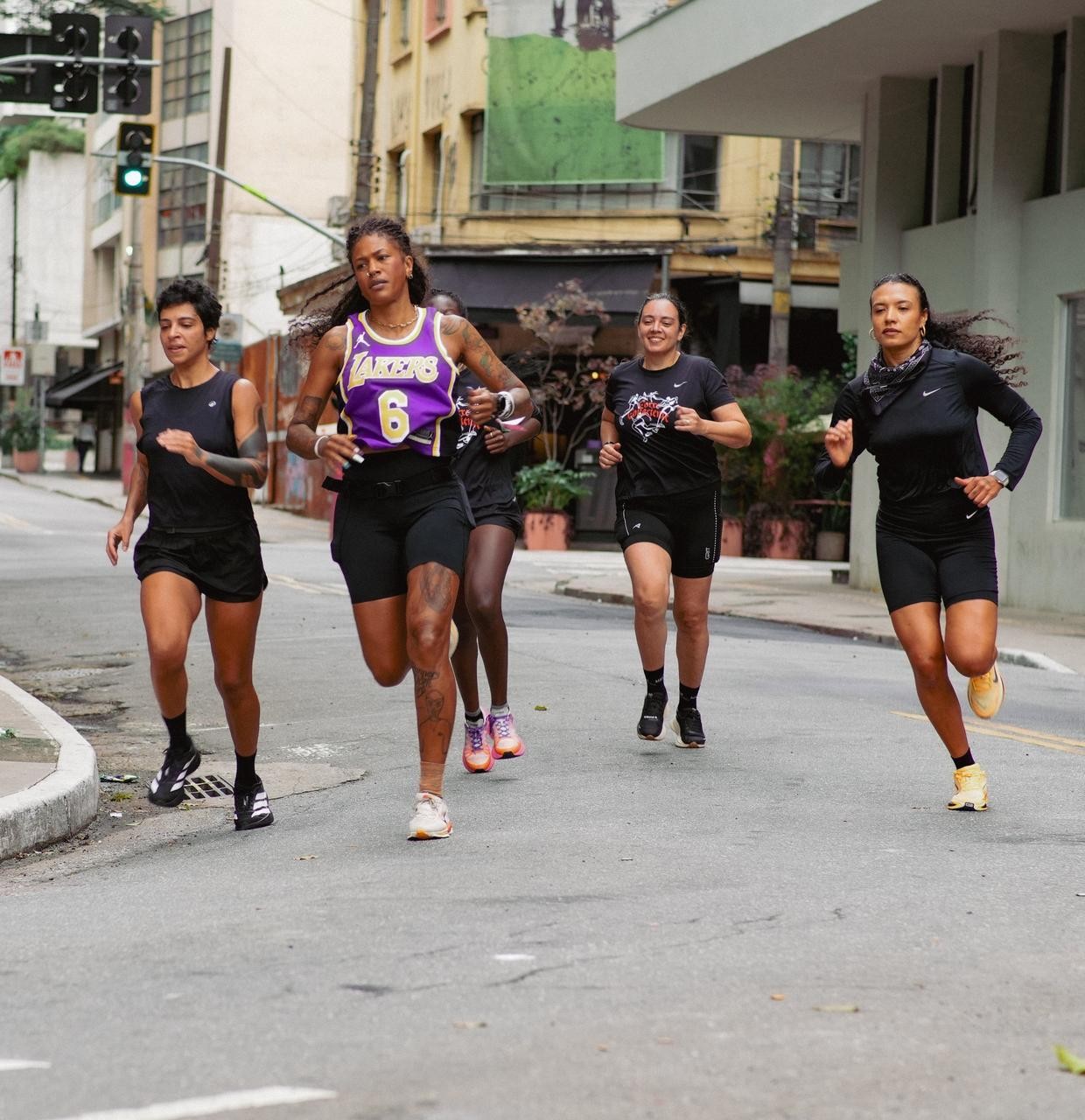 Cinco mulheres correndo na rua, a primeira veste regata roxa e amarela dos Lakers, as outras usam roupas esportivas pretas. Elas est&atilde;o em uma rua urbana com pr&eacute;dios antigos ao fundo e um sem&aacute;foro verde