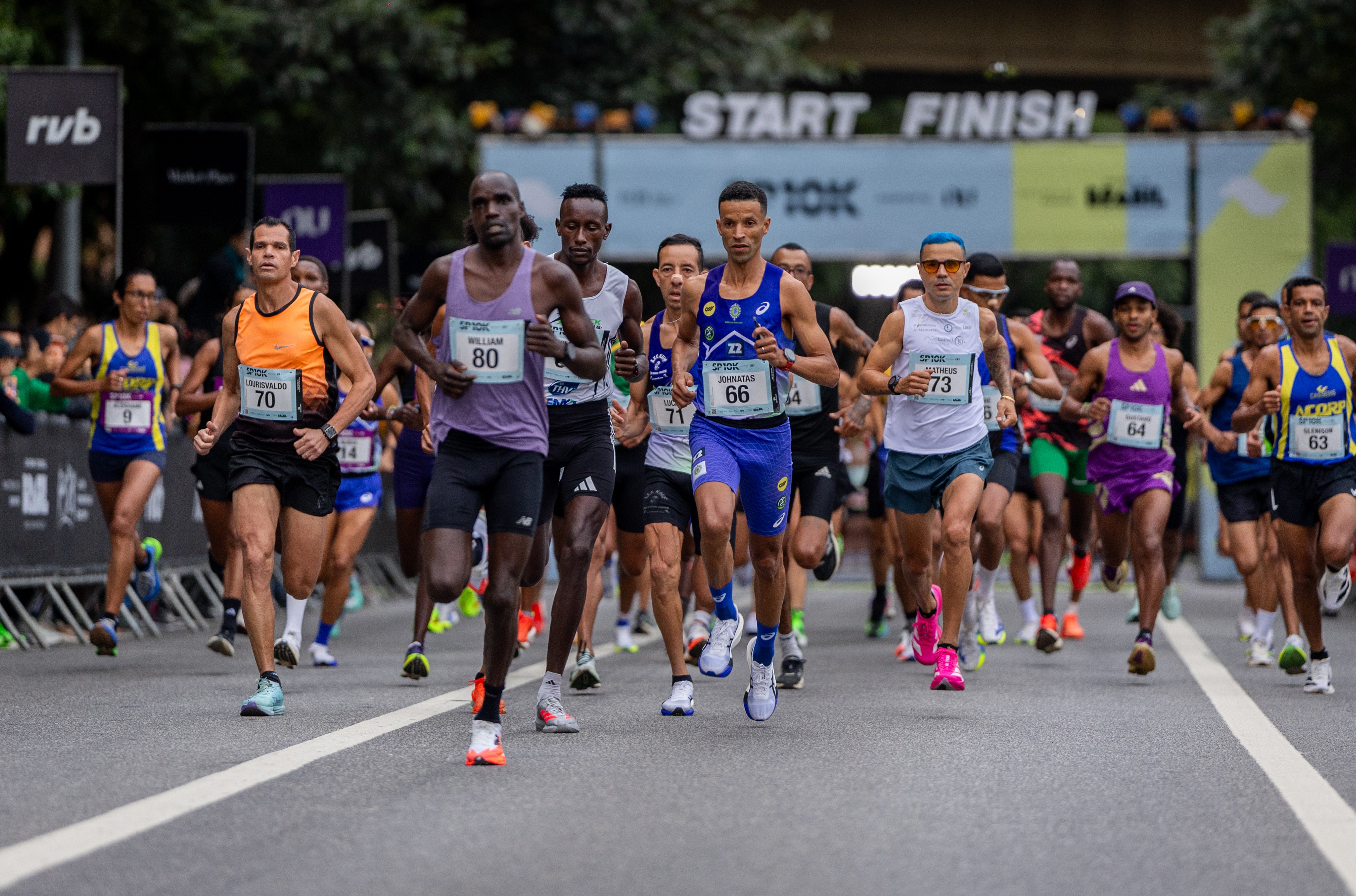 Corredores de diversas etnias e idades participam de uma maratona, vestindo regatas e shorts coloridos com n&uacute;meros de identifica&ccedil;&atilde;o. Eles correm em uma rua asfaltada, com uma faixa branca central, sob um p&oacute;rtico 