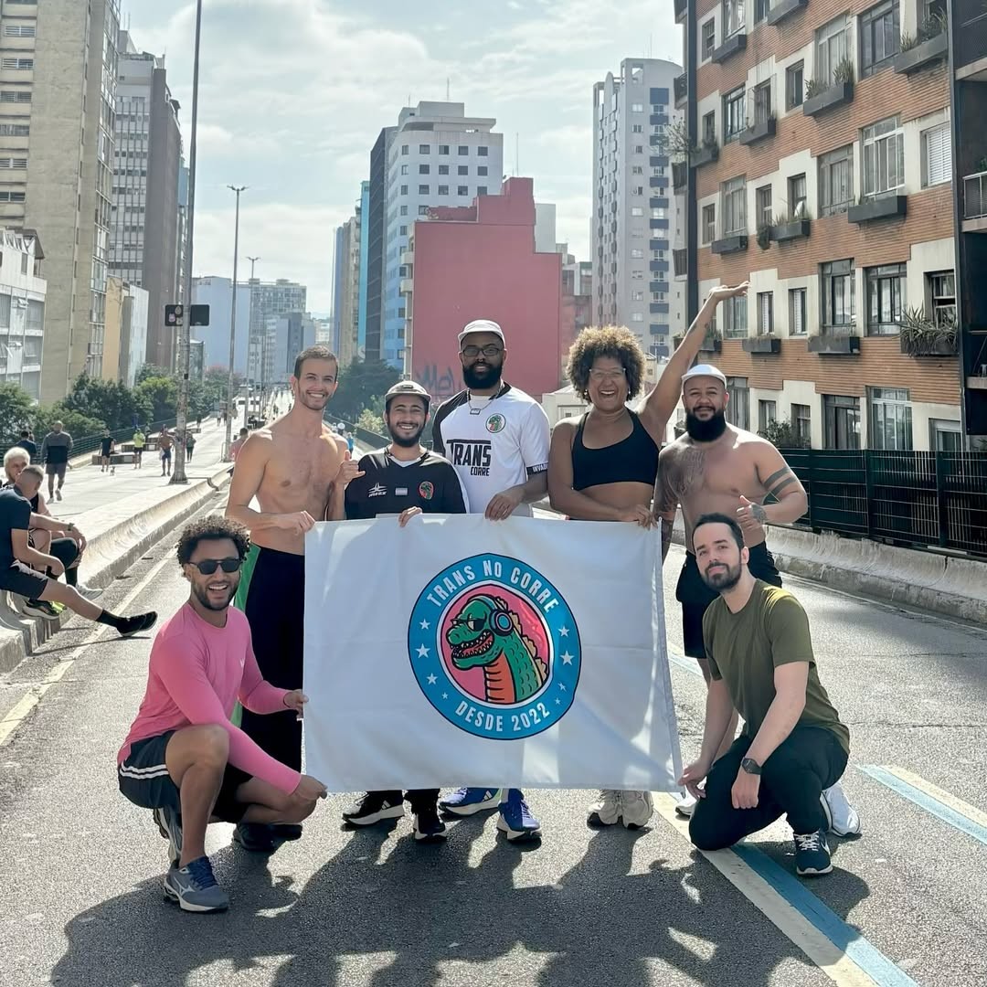 Sete pessoas sorridentes, algumas sem camisa, seguram uma bandeira branca com o logo Trans no Corre Desde 2022 em uma rua urbana com pr&eacute;dios altos ao fundo