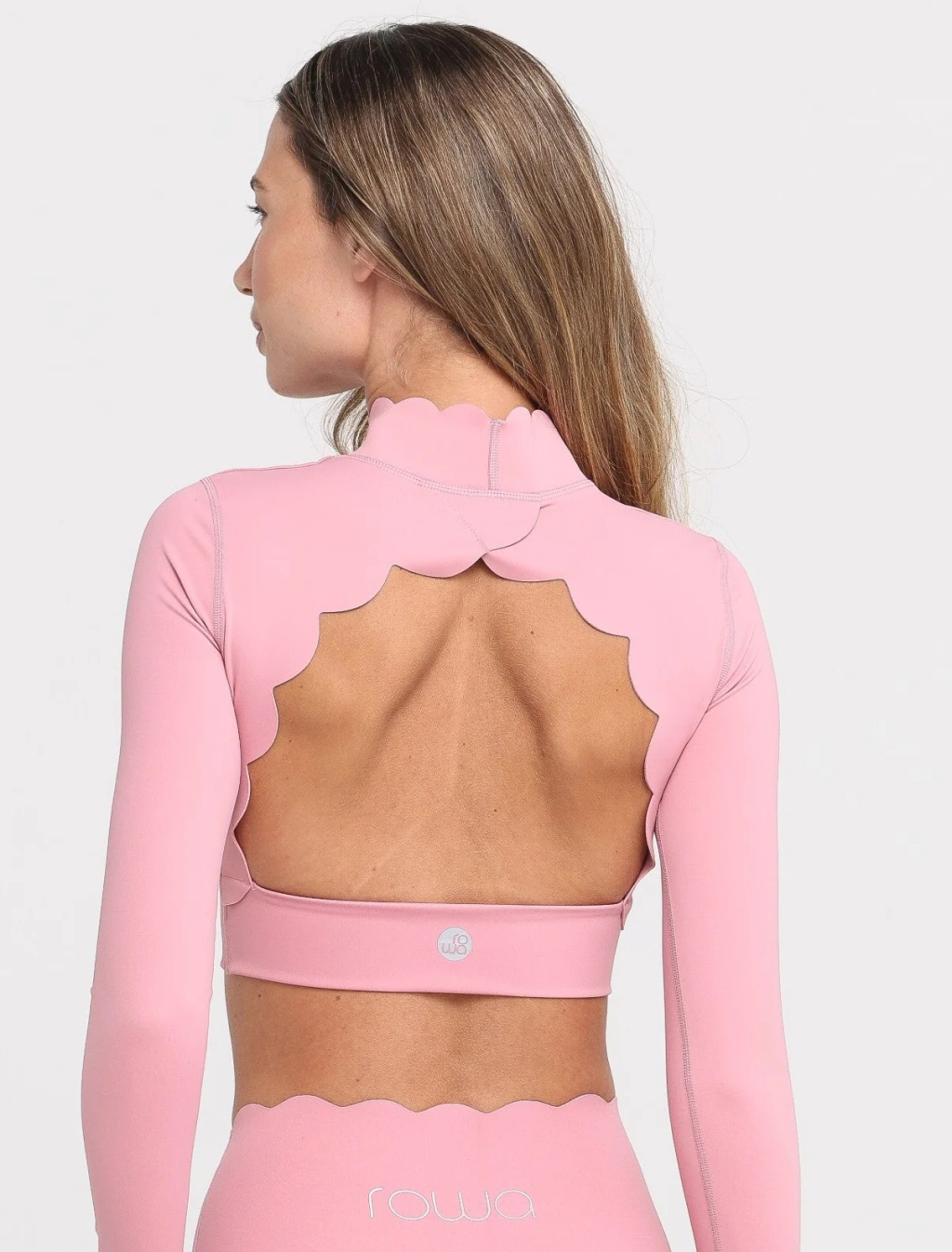 Mulher de costas, com cabelos castanhos longos, vestindo um conjunto esportivo rosa claro. A blusa de manga comprida tem gola alta e recortes ondulados nas costas, expondo a parte superior das costas. A cal&ccedil;a tem cintura alta e acabamento ondulado na borda superior. O logo rowa aparece na cal&ccedil;a e um s&iacute;mbolo circular na blusa