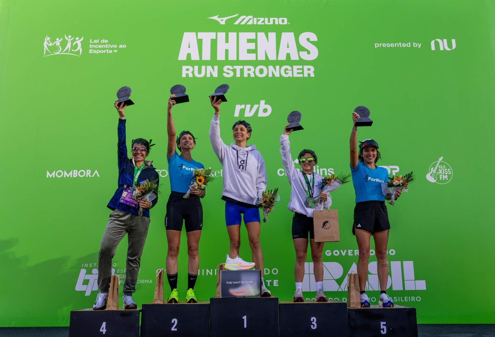 Cinco mulheres atletas sorridentes no p&oacute;dio de uma corrida, levantando trof&eacute;us e segurando buqu&ecirc;s de girass&oacute;is, em frente a um painel verde com logos de patrocinadores