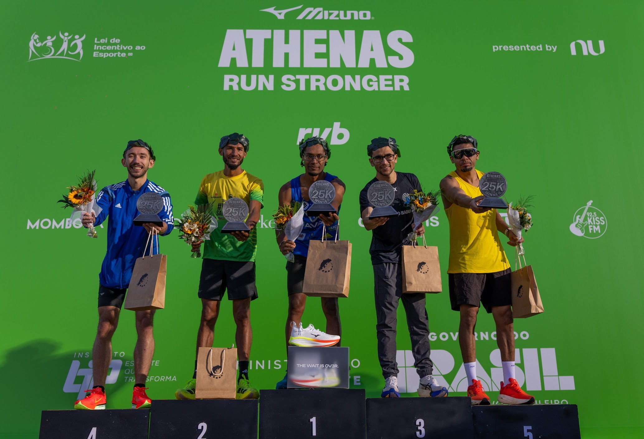 Cinco homens sorridentes, usando coroas de louros, em um p&oacute;dio de corrida. O primeiro lugar exibe um t&ecirc;nis branco, os outros seguram trof&eacute;us, buqu&ecirc;s e sacolas de papel. O fundo verde tem o logo ATHENAS RUN STRONGER e patrocinadores
