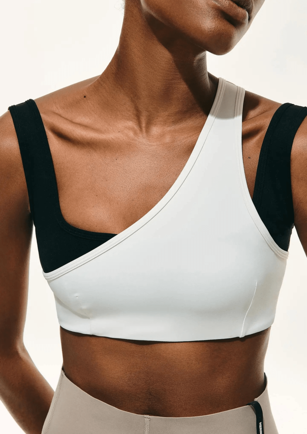 Mulher com pele escura usando um top esportivo branco de al&ccedil;a &uacute;nica sobre um top preto, e cal&ccedil;a legging bege. A imagem foca no tronco e pesco&ccedil;o da modelo, com ilumina&ccedil;&atilde;o forte e fundo branco
