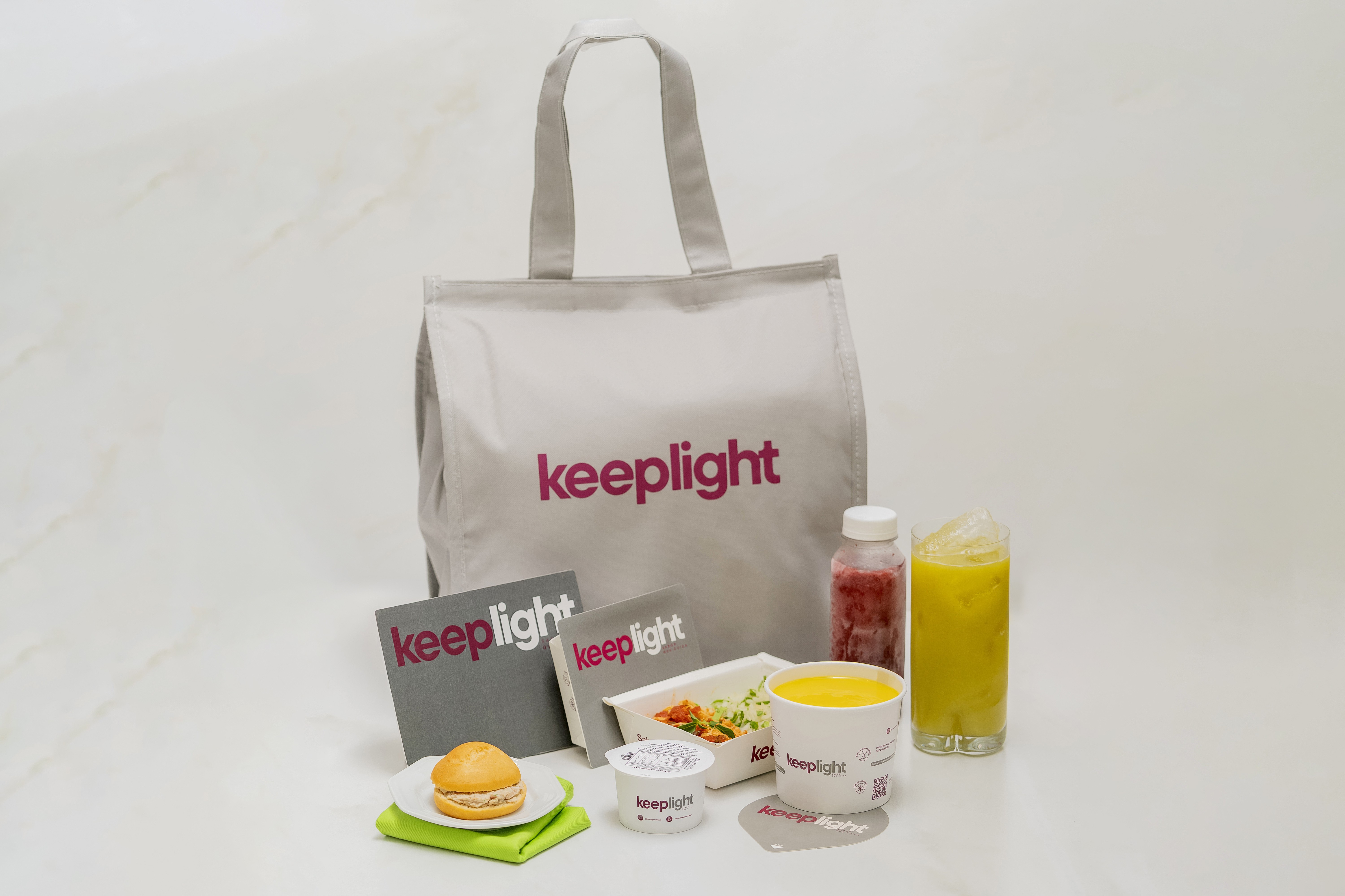 Uma sacola t&eacute;rmica cinza com o logo keeplight em rosa, acompanhada de refei&ccedil;&otilde;es e bebidas saud&aacute;veis. H&aacute; um sandu&iacute;che em prato branco, dois folhetos, um pote de iogurte, uma marmita, um copo de sopa, uma garrafa de suco vermelho e um copo de suco amarelo com gelo, todos com a marca keeplight