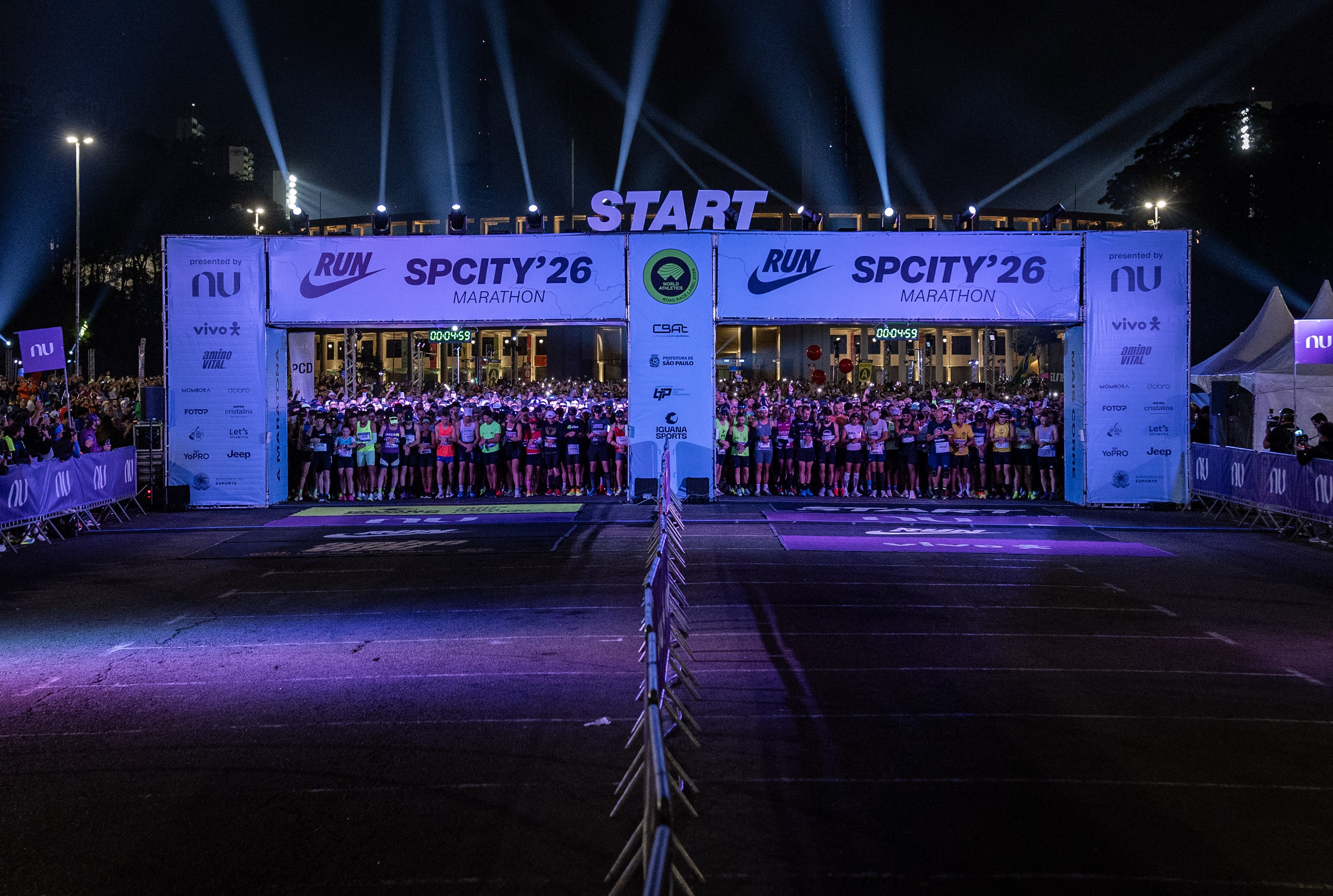 Corredores aguardam o in&iacute;cio da SP City Marathon 26 sob um portal START iluminado por holofotes, com banners de patrocinadores e um cron&ocirc;metro digital exibindo 00:04:59. A multid&atilde;o, vestindo roupas esportivas, est&aacute; alinhada na rua escura, que reflete a ilumina&ccedil;&atilde;o roxa do evento.