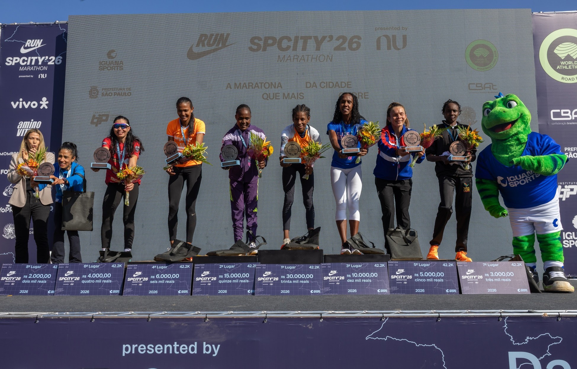 Oito mulheres e um mascote verde em um p&oacute;dio de premia&ccedil;&atilde;o de maratona. As mulheres seguram trof&eacute;us e buqu&ecirc;s, vestindo roupas esportivas coloridas. O mascote, um lagarto verde com camiseta azul Iguana Sports, est&aacute; &agrave; direita. No fundo, um tel&atilde;o exibe logotipos de patrocinadores e informa&ccedil;&otilde;es da SPCITY26 Marathon. Abaixo do p&oacute;dio, placas indicam os lugares, valores de premia&ccedil;&atilde;o e dist&acirc;ncia de 42.2K