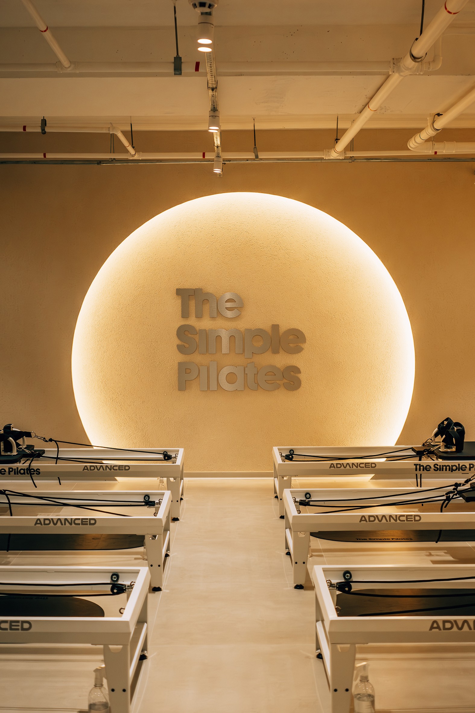 Est&uacute;dio de Pilates moderno com paredes em tom bege claro e teto branco. No centro, uma parede com um c&iacute;rculo iluminado por tr&aacute;s, onde se l&ecirc; The Simple Pilates em letras met&aacute;licas. Seis m&aacute;quinas de Pilates brancas com detalhes pretos e a palavra ADVANCED em destaque est&atilde;o dispostas em duas fileiras, tr&ecirc;s de cada lado, no ch&atilde;o de azulejos claros. Garrafas de &aacute;gua e borrifadores de limpeza s&atilde;o vis&iacute;veis ao lado das m&aacute;quinas.