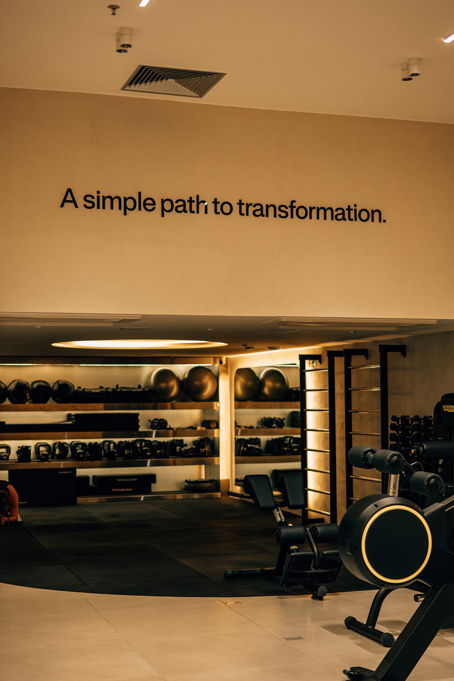 Academia moderna com piso de borracha e azulejos claros, prateleiras com halteres, kettlebells e bolas de gin&aacute;stica. Na parede bege, a frase A simple path to transformation em preto. Equipamentos de muscula&ccedil;&atilde;o escuros com detalhes dourados est&atilde;o &agrave; direita