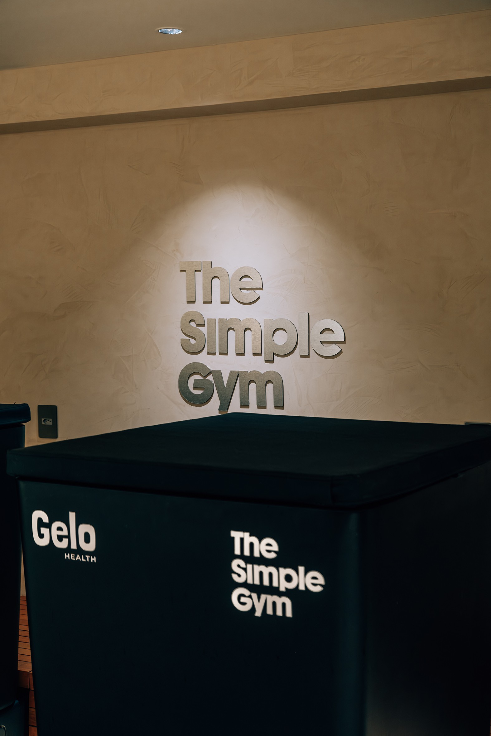 O letreiro The Simple Gym em relevo e iluminado na parede bege texturizada, acima de um balc&atilde;o preto com os logotipos Gelo HEALTH e The Simple Gym em branco
